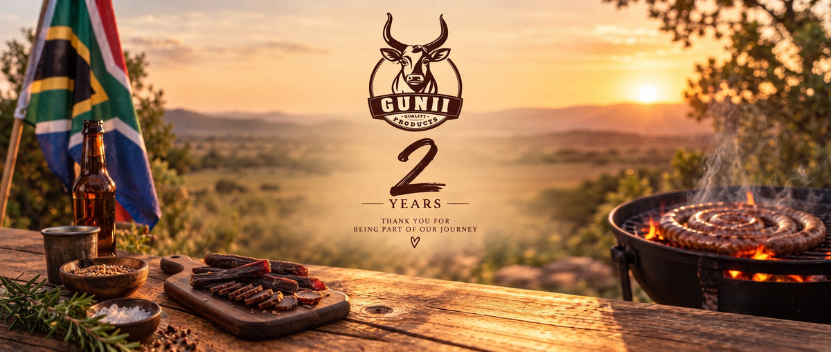 Outdoor sunset scene with a grill/Braai, Boerewors cooking, a wooden cutting board with Biltong and Droewors, a bottle, bowls, and a South African flag on a table. A large central logo reads 'GUNII 2 YEARS THANK YOU FOR BEING PART OF OUR JOURNEY'.
